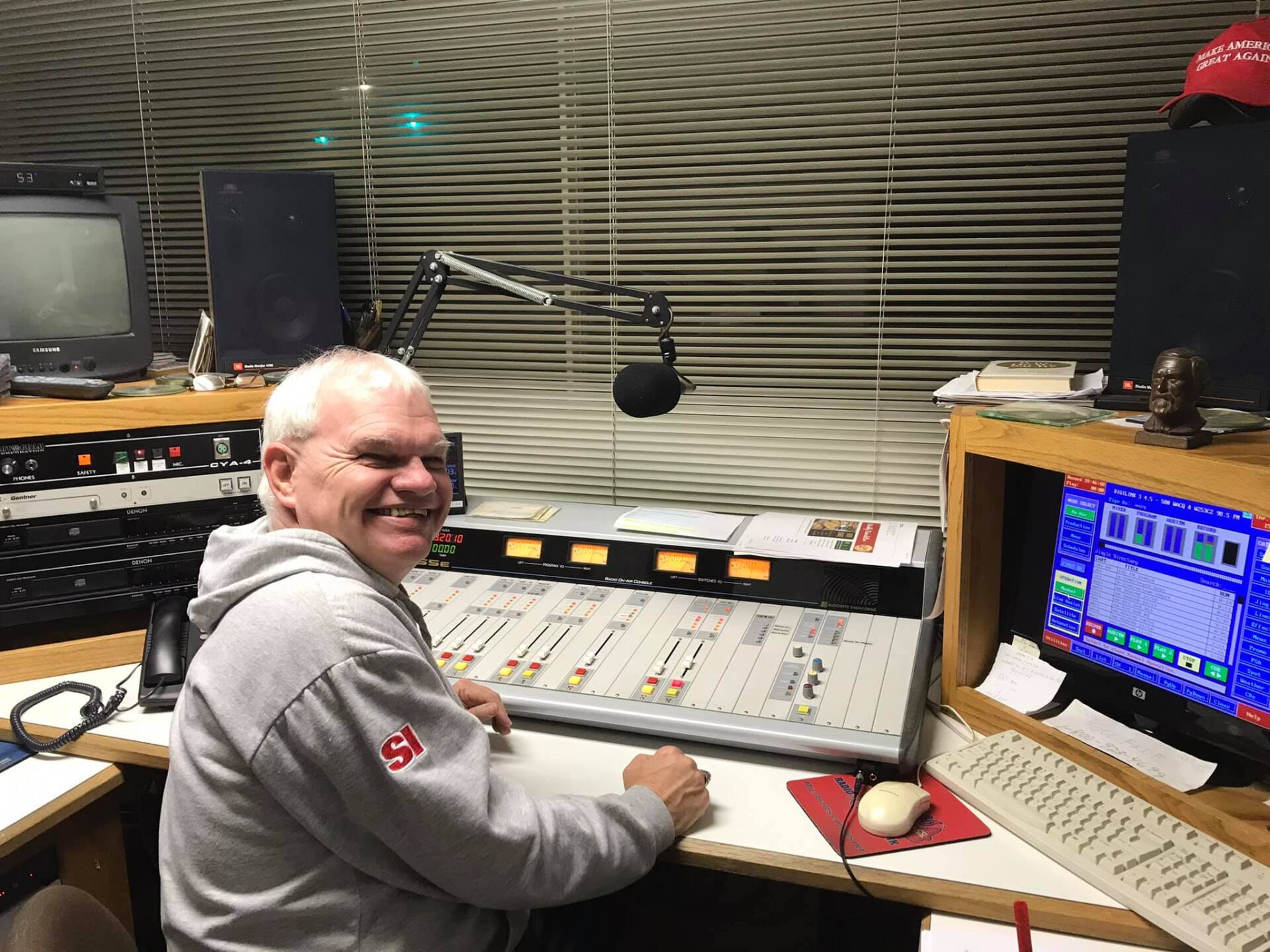 Man in radio studio smiling at the camera, seated at a soundboard.