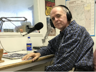 Man in plaid shirt wearing headphones at a radio desk with a microphone and water bottle.