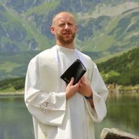 Man in white robe holds a book, standing near a lake with mountains in the background.
