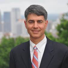 Headshot of a man in a dark suit and striped tie, smiling with a blurred city skyline behind him