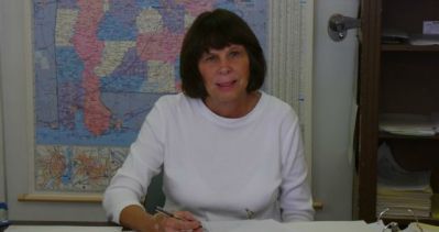 Woman in white shirt, seated at a desk, facing the camera, with a map in the background.