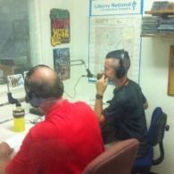 Two men in a radio studio at desks, wearing headsets, with microphones. One in red shirt, one in black.