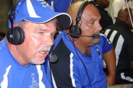 Two men in blue jerseys and headsets sitting closely together, likely broadcasting at a sports event.