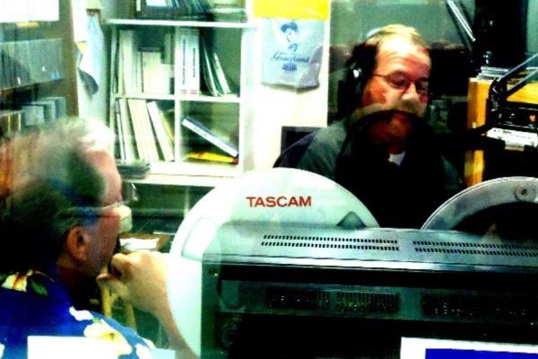 Two men in a radio studio. One with a large mustache and headset, operates equipment. The other listens.