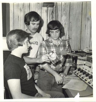 Three people sit at a control panel in a wood-paneled room, examining a handheld device.