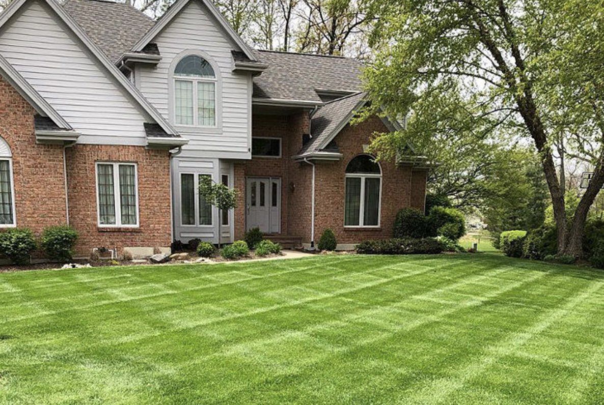 Brick house with a manicured lawn featuring diagonal stripes, trees and landscaping in the yard.