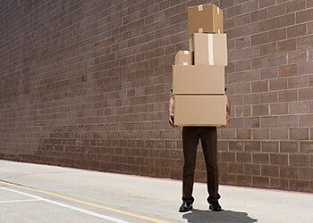 Person holding a stack of cardboard boxes in front of a brick wall, standing on pavement.