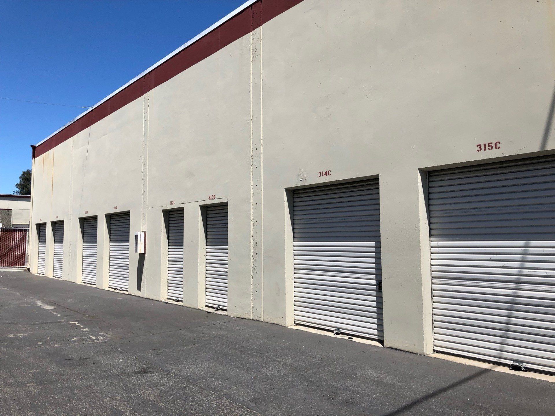 Storage units in a row, with closed white doors and tan walls under a red roof, on a paved lot.