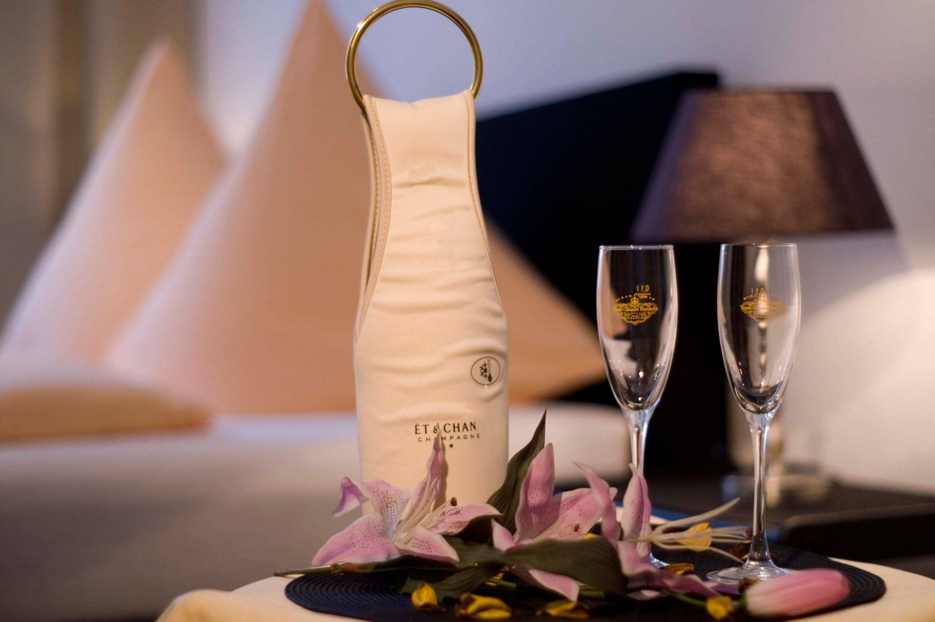 Eine Champagnerflasche in einer Schutzhülle, dazu zwei Sektgläser, rosa Lilien und eine Lampe auf einem Nachttisch.
