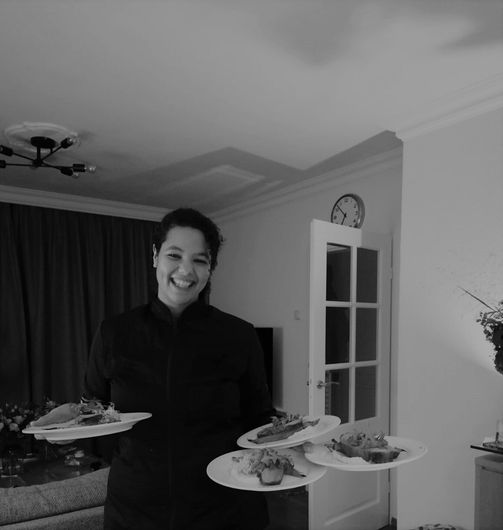Een glimlachende persoon in een zwart koksuniform houdt vier borden met eten vast in een eetkamer.