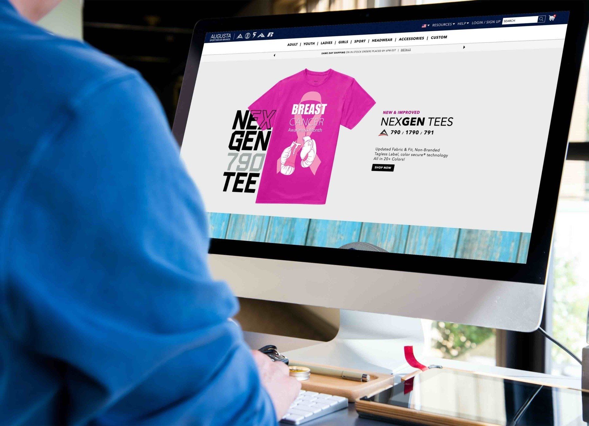 A person in a blue shirt works at a computer displaying a website for custom pink awareness t-shirts.