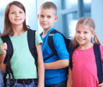 Three children with backpacks are standing next to each other in a hallway.