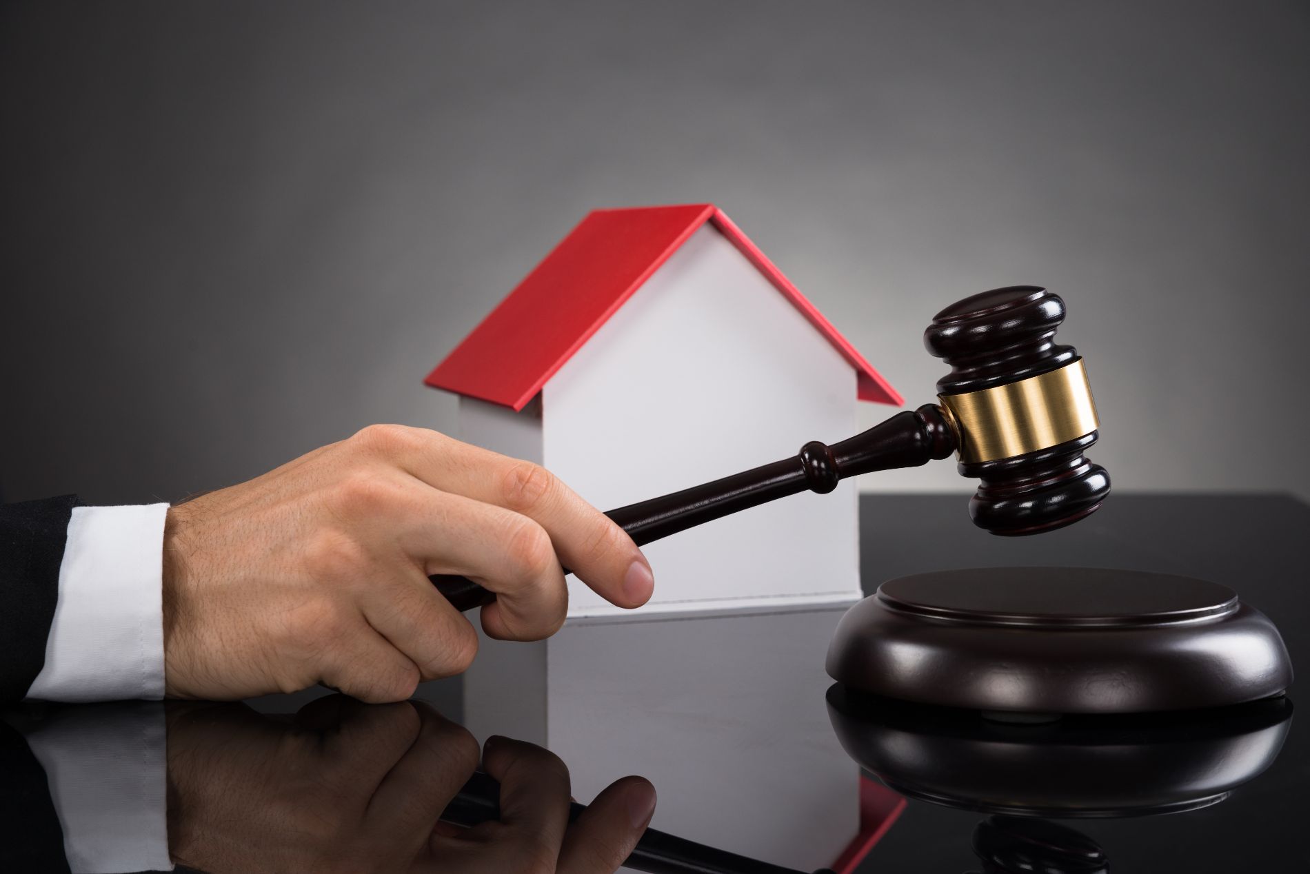 Hand holding gavel about to strike against a base, with a model house in the background.