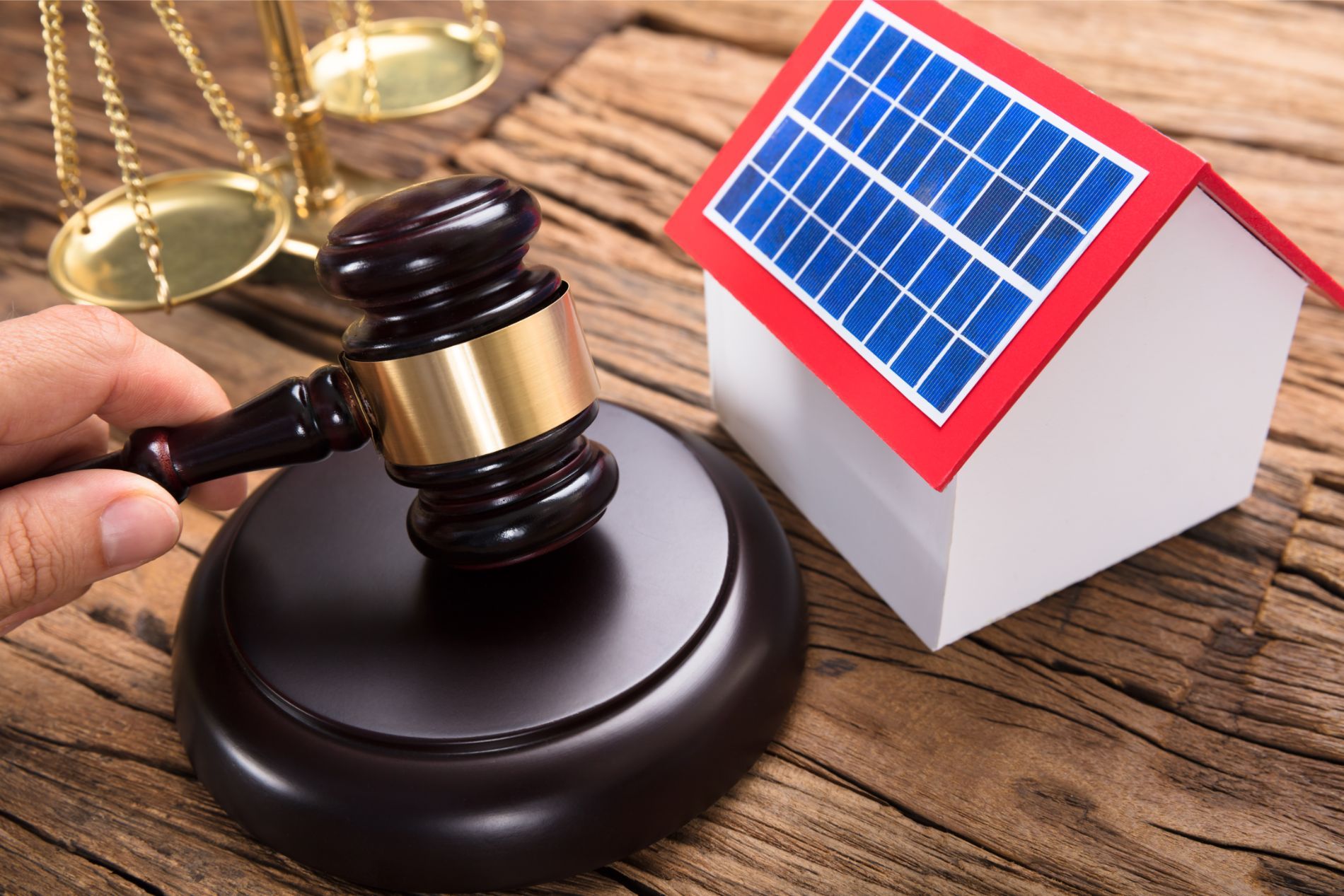 Hand holding gavel next to miniature house with solar panels, scales of justice on wood.
