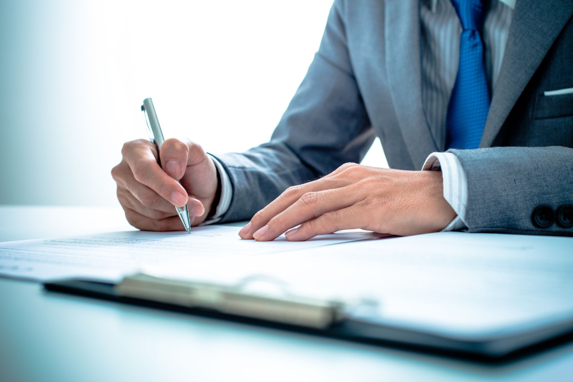 Man in a suit signing paperwork at a desk; pen in hand, clipboard in foreground.