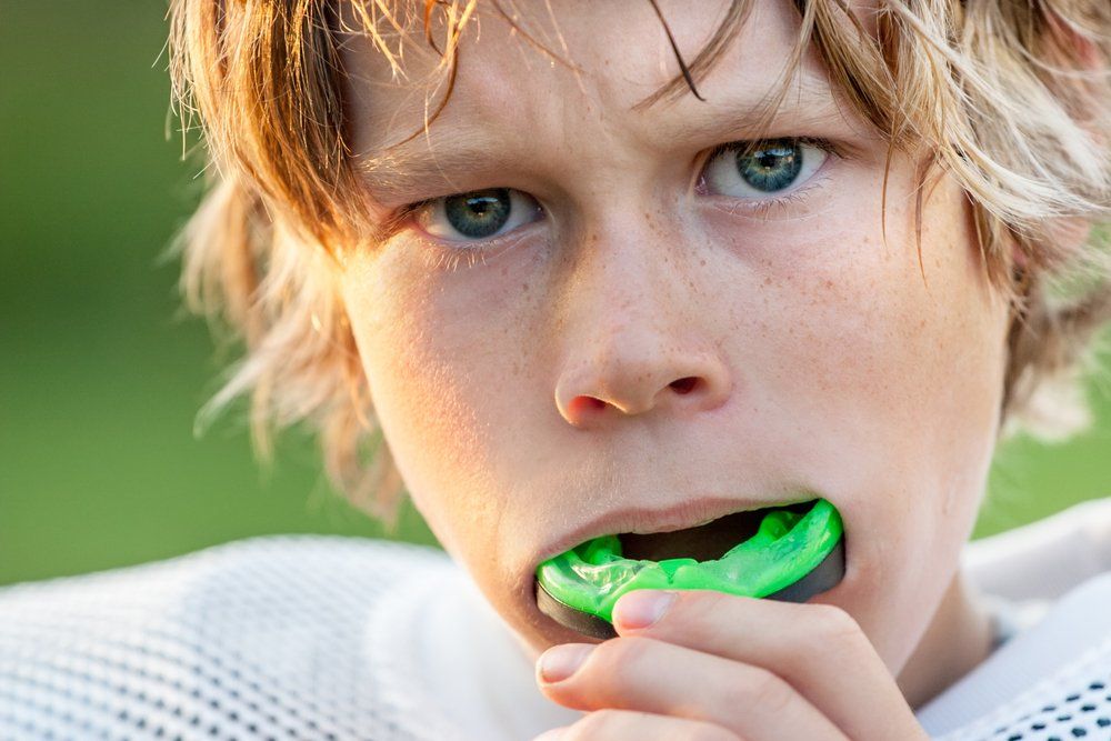 Boy with blonde hair, wearing a green mouthguard, holding it in his mouth, looking intensely at the camera.