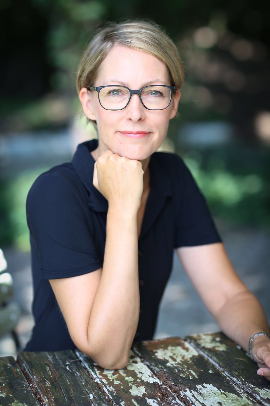 Woman with glasses resting chin on hand, sitting at a weathered table outside.