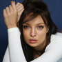 Woman with brown hair, in white top, leans forward with hands on her head, looking at the viewer against a blue backdrop.