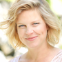 Woman with blonde hair smiles, blurred outdoor background.