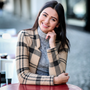Woman smiling, wearing a plaid blazer and gray turtleneck, leaning on a table outdoors.