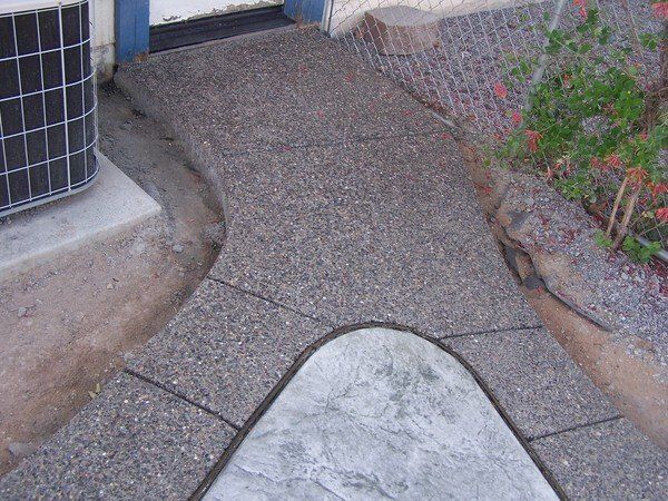 A concrete walkway leading to a house with flowers in the background