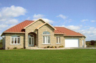 Maison en pierre beige avec toit rouge, porte de garage blanche et pelouse verte sous un ciel bleu.