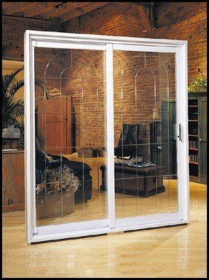 Porte coulissante en verre à cadre blanc avec croisillons décoratifs. Vue intérieure : salon meublé, mur en briques apparentes.