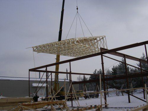 Une grue soulève une ferme de toit en bois au-dessus d'une structure métallique sur un chantier de construction recouvert de neige.