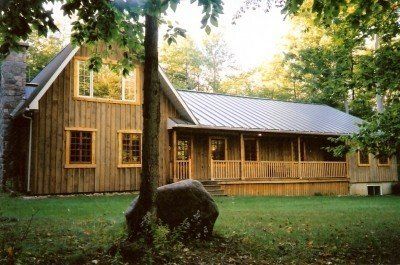 Cabane en bois avec véranda, cheminée en pierre et cour verdoyante entourée d'arbres.