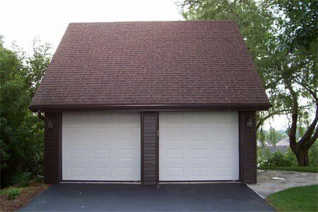 Garage double au toit brun et portes blanches, donnant sur une allée en asphalte noir. Arbres en arrière-plan.