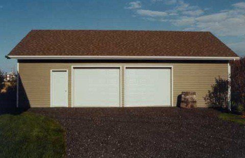 Garage beige pour deux voitures, avec un toit marron et une porte latérale, donnant sur une allée de gravier sous un ciel bleu.