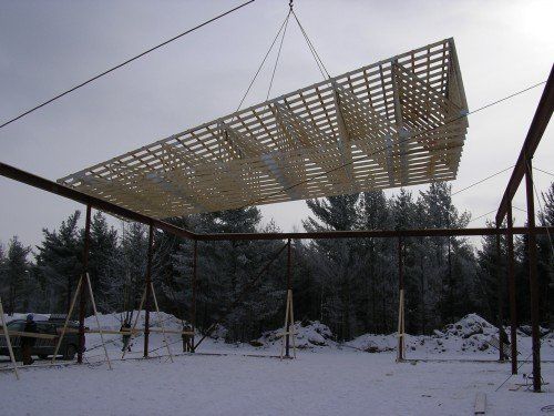 Une structure en bois est soulevée par une grue pour être placée sur une structure en acier dans un décor extérieur enneigé.