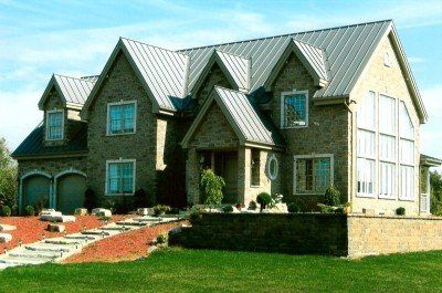 Maison en briques avec toit en métal, plusieurs lucarnes, fenêtres cintrées et aménagement paysager soigné.