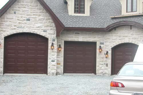 Portes de garage marron dans un bâtiment en pierre, sous un toit gris. Voiture garée à droite.