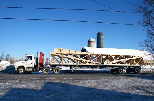 Camion transportant une grande charpente en bois sur une route enneigée, avec des silos et un bâtiment en arrière-plan.