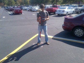 Person in parking lot holding surveying equipment, looking at a device. Cars are parked nearby.