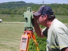 Surveyor using a theodolite on a tripod outdoors, green field and trees in the background.