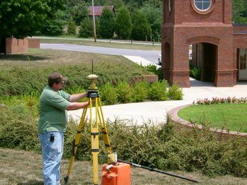 Man using surveying equipment on a tripod near a brick building.