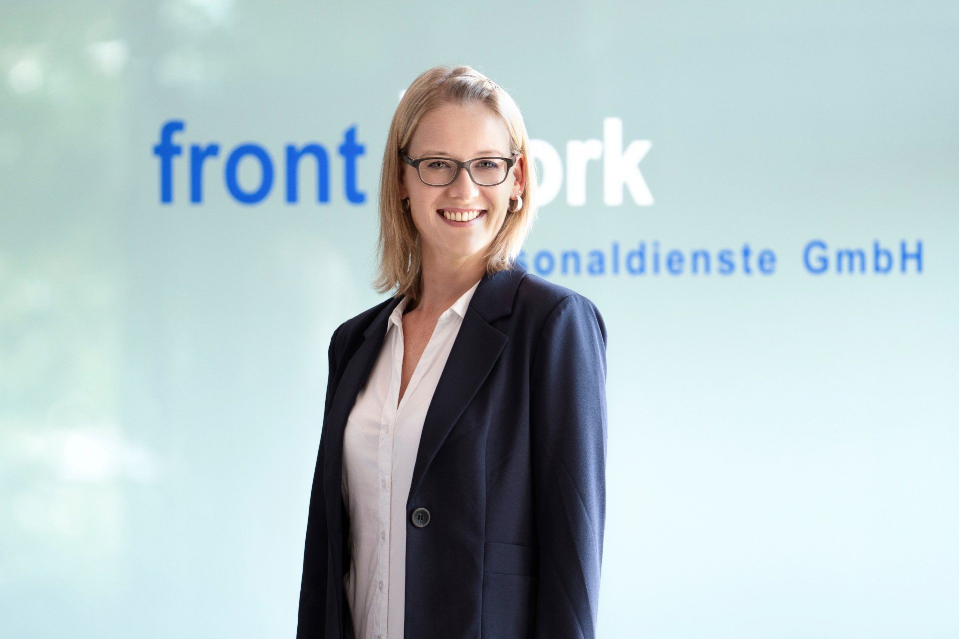Eine Frau im Anzug und mit Brille steht vor einem Schild mit der Aufschrift „Front Work“.