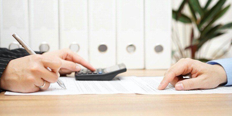Two hands writing on documents at a desk with a calculator and binders nearby