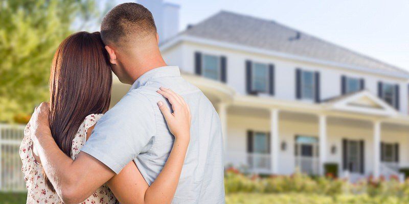 Couple embracing in front of a white house with a porch and green shutters