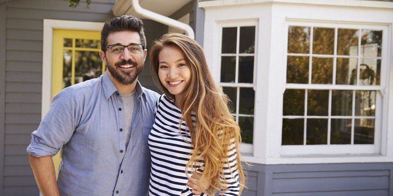 Smiling couple standing outside a house, with a man in a blue shirt and a woman in a striped top.