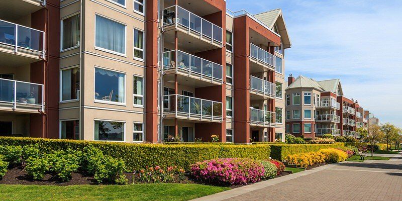 Modern apartment buildings with balconies, landscaped flower beds, and a tree-lined walkway