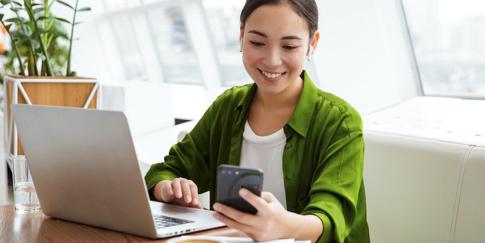 Smiling person in a green shirt using a laptop and phone on a couch.