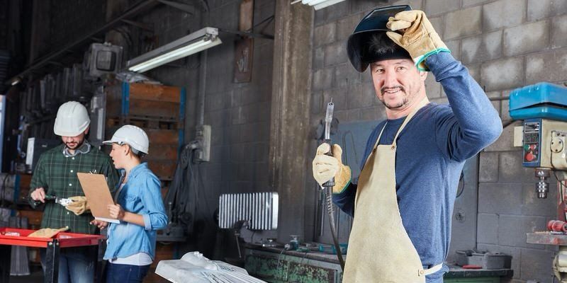 Workers in a workshop, one smiling man in an apron and gloves holding up a part.
