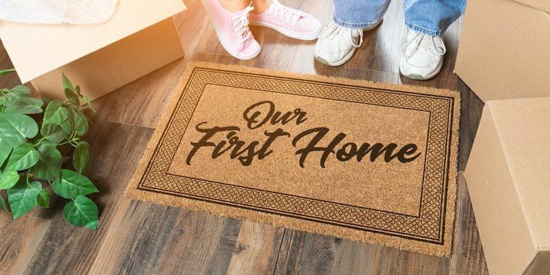 Welcome-home doormat reading “Our First Home” in an open cardboard box