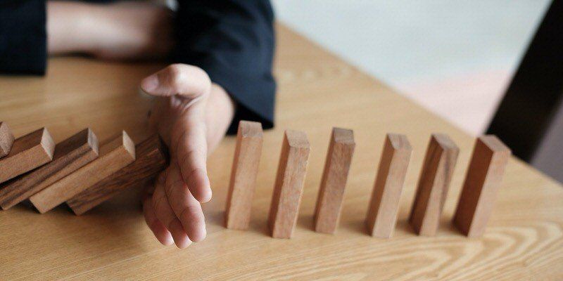 Hand placing a wooden block in a row of dominoes on a table, suggesting a chain reaction
