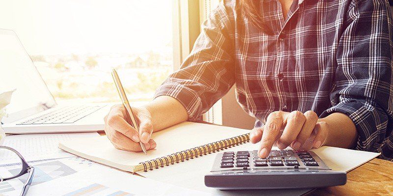 Person writing in a notebook beside a calculator at a sunlit desk, reviewing paperwork