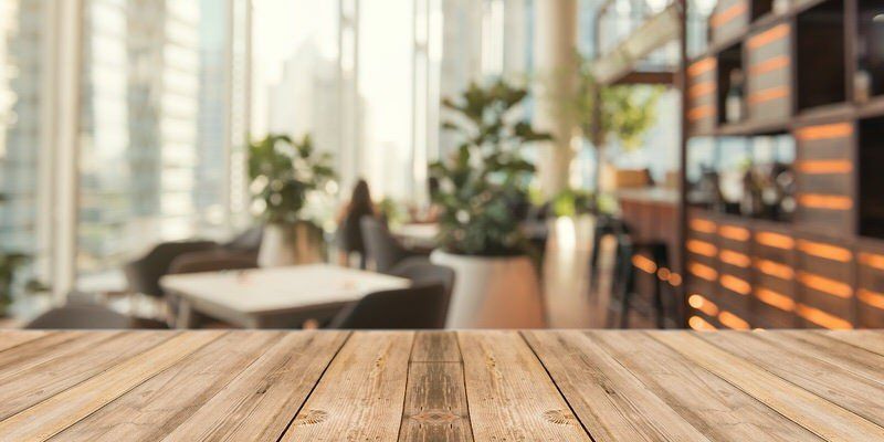 Wooden tabletop in a bright cafe with blurred chairs, plants, and large windows in the background