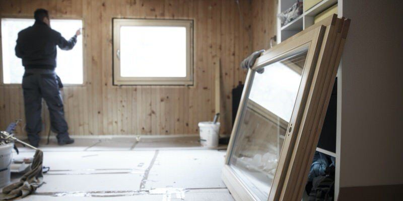 Person installing wall paneling in a room with wood-paneled walls and a window, construction materials on the floor
